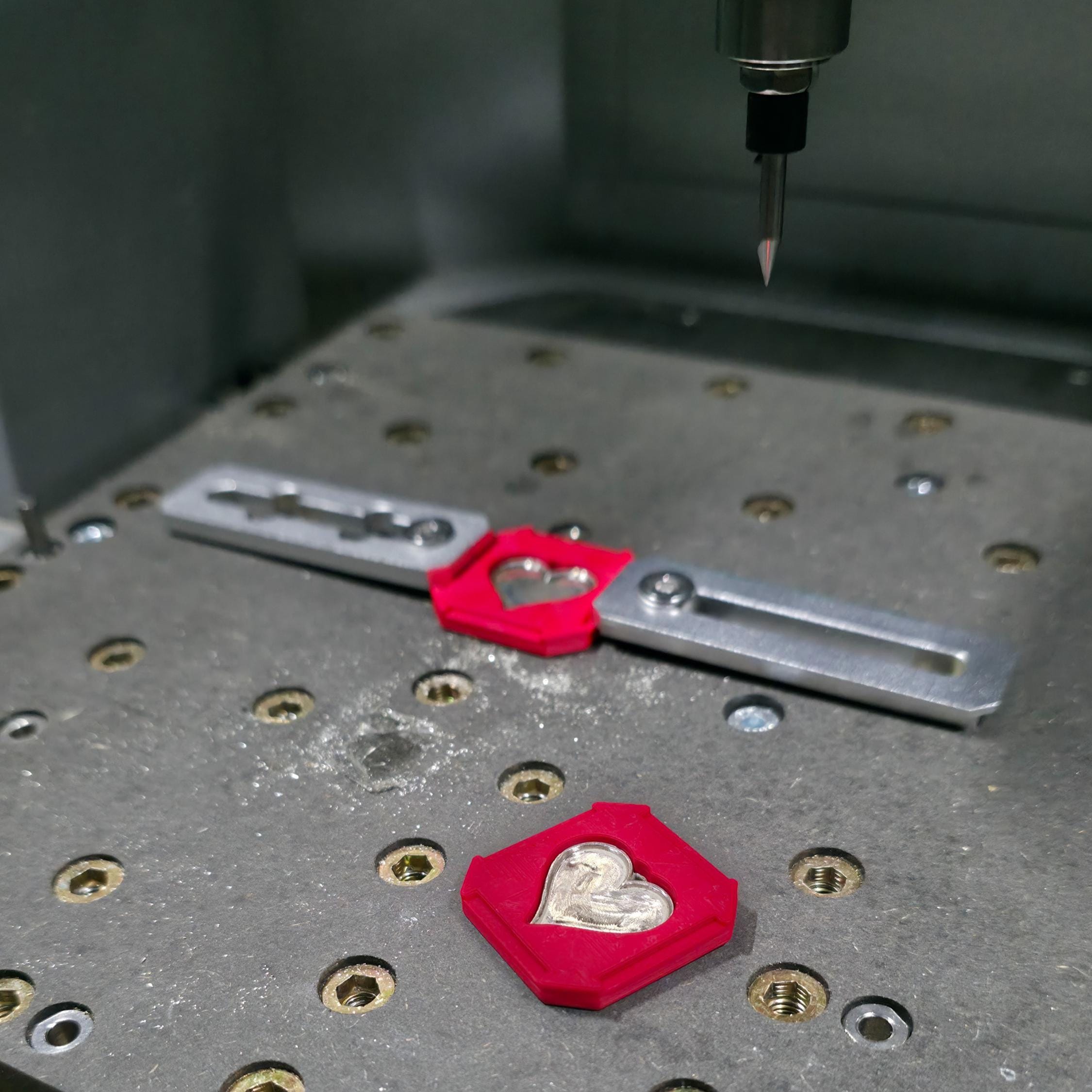 “Close-up of a desktop CNC milling machine bed showing a precision engraving setup for custom jewelry. A small carbide bit hangs above a metal fixture holding a red 3D-printed jig with a heart-shaped silver pendant blank, with another matching jig and heart on the perforated work table, illustrating prototype machining for personalized jewelry parts.”
