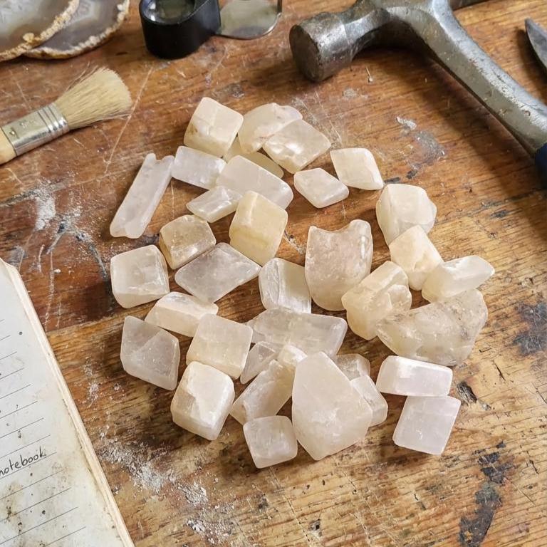 Lightly tumbled calcite chips arranged on a dusty workshop bench surrounded by mineral specimens, tools, sample containers, and a jeweler’s loupe. The chips are pale cream to light yellow with clean edges. Scene evokes a working lapidary or mineral-prep studio, highlighting the material’s readiness for shaping, cutting practice, or educational demonstration.