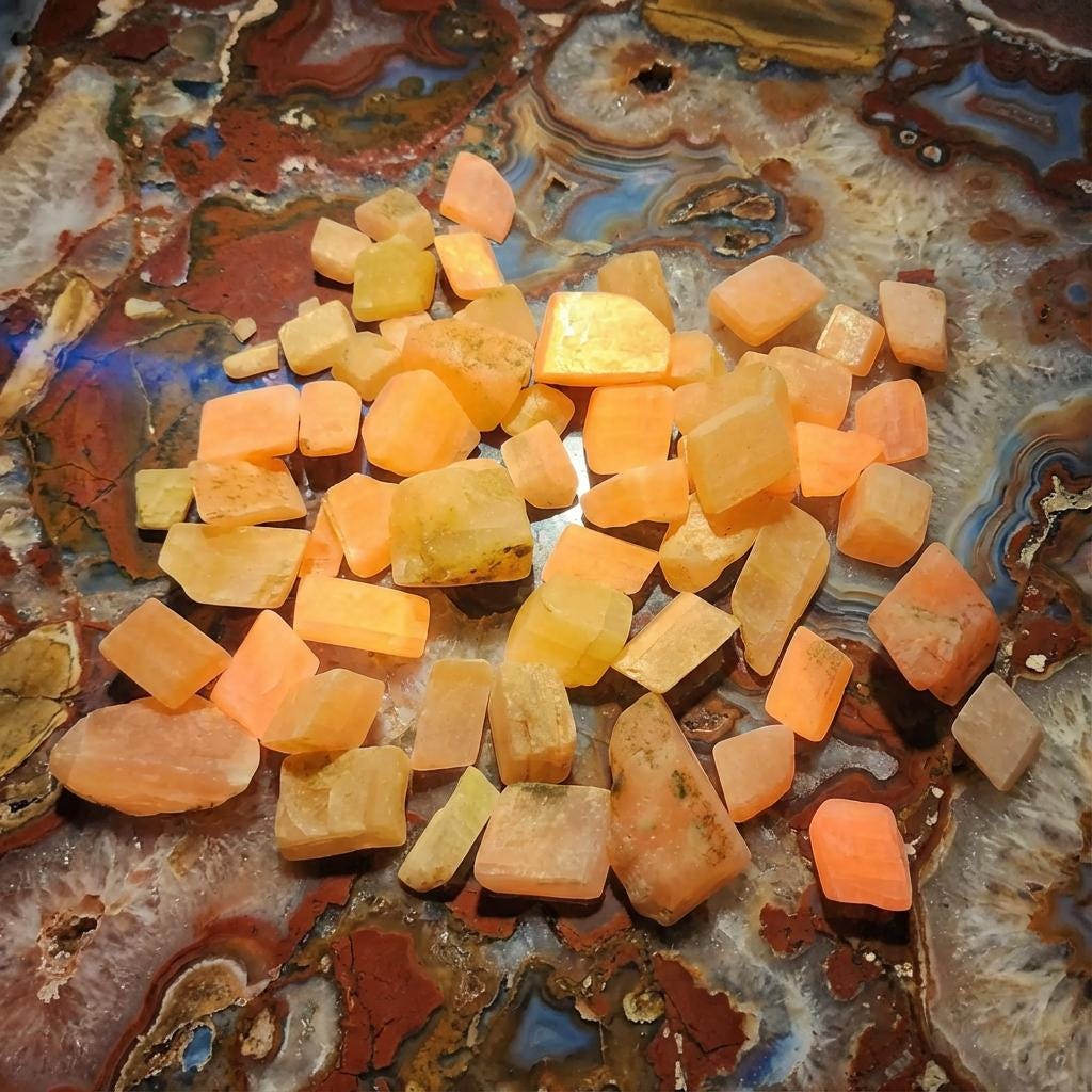 A spread of lightly tumbled fluorescent calcite chips on natural rock slabs surrounded by field tools and mineral specimens. The calcite pieces display mixed orange, yellow, and cream coloration with clean tumbled edges. Scene evokes rockhounding and geological study, showing the material in a natural field-collection context for lapidary, inlay, and teaching applications.