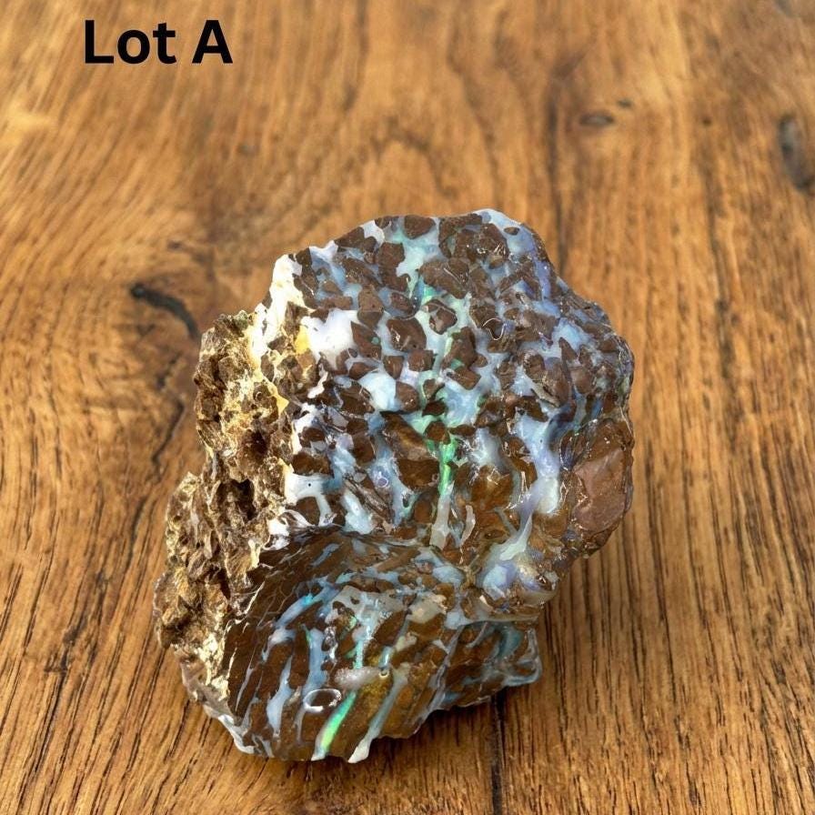 Australian Boulder Opal rough labeled Lot A, sitting on a wooden surface. The stone has thick blue and green opal veins woven through brown ironstone with patches of white host material. Strong fire visible in natural cavities. Suitable for cabbing, contour shaping, and lapidary orientation work. Untreated Queensland opal.