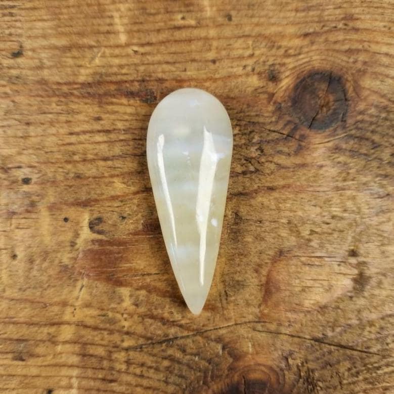 Polished teardrop-shaped chalcedony agate cabochon resting on textured wood. The stone shows soft milky translucence with pale yellow particulate inclusions from ancient volcanic sands. Subtle banding and internal haze are visible under directional light, highlighting the Oligocene-age silica structure typical of Pacific Northwest agates.