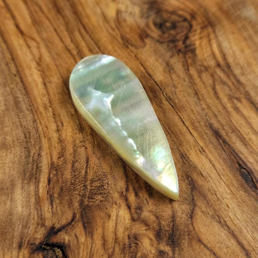 Angled upside down close-up of a polished teardrop chalcedony agate on warm wood. Internal yellow and green tones brighten under the light, revealing embedded volcanic particulates and soft silica banding. The cabochon’s curved surface and natural inclusions show clear Oligocene-age chalcedony structure.