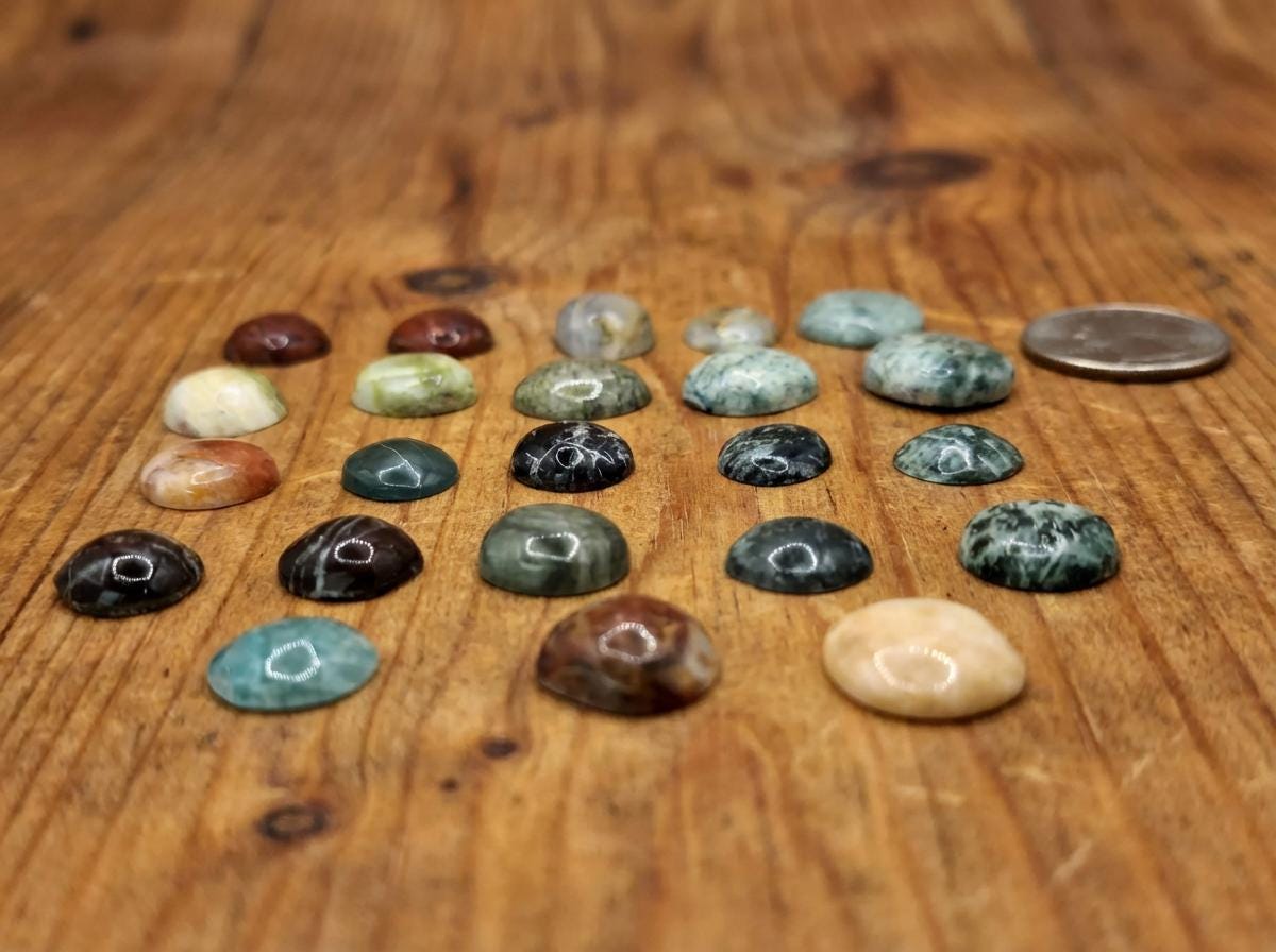 Side-angle view of cabochon gemstones arranged on a rustic wood surface. The domed stones include green jasper, black with white veining, red-brown patterns, and teal chalcedony. The low camera angle emphasizes dome height and polish quality.