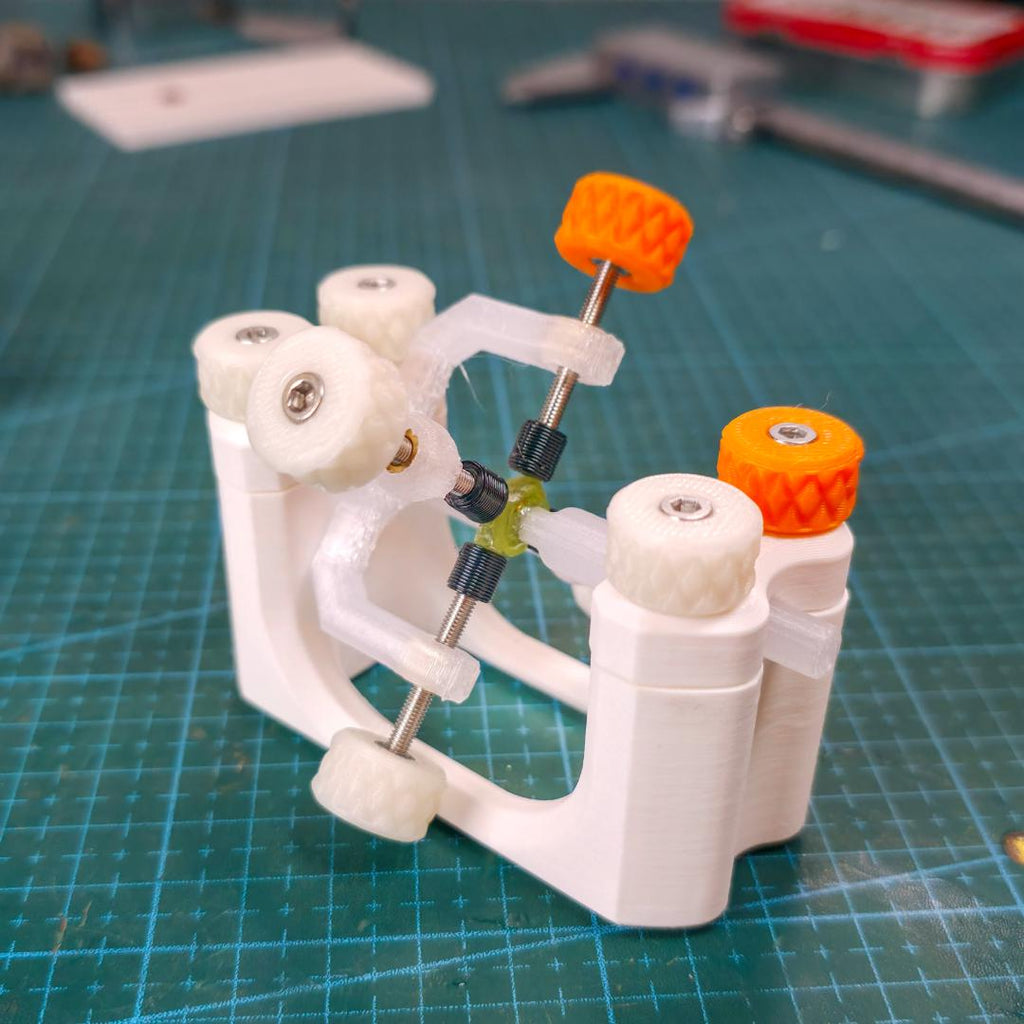“Close-up of the Gem-Xfer epoxy cure station with a faceting transfer jig mounted in the frame, showing the adjustable M3 screw system, translucent arms, and bioplastic knurled knobs. The dop and stone are centered in the curing position on a green cutting mat. Tools and lapidary gear sit in the background, emphasizing makerspace use.”