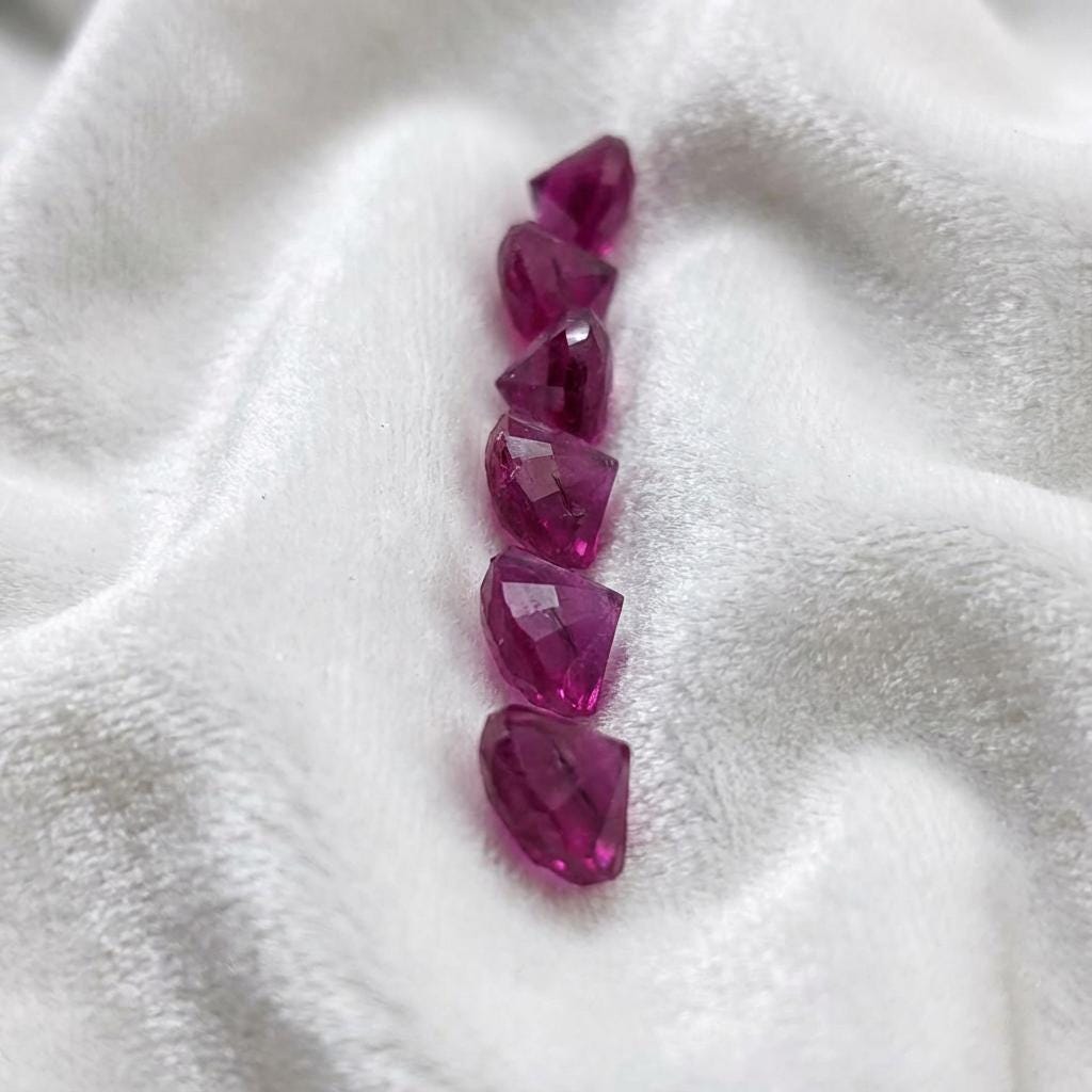 Side profile view of lab grown pink sapphire stones, 2 mm round cut, arranged in a vertical line on white fabric. Pavilion depth and facet structure visible, showing consistent cut quality for use as accent stones in custom jewelry and metalwork.