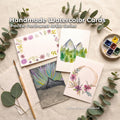 “Handmade watercolor card featuring a Pacific Northwest mountain peak with aurora-inspired rays in soft greens, violets, and gold. The card rests on a warm wooden table, highlighting the textured watercolor paper. Part of the Aetherium x Archibald PNW Artist Series, created as a small-batch handmade card set.”