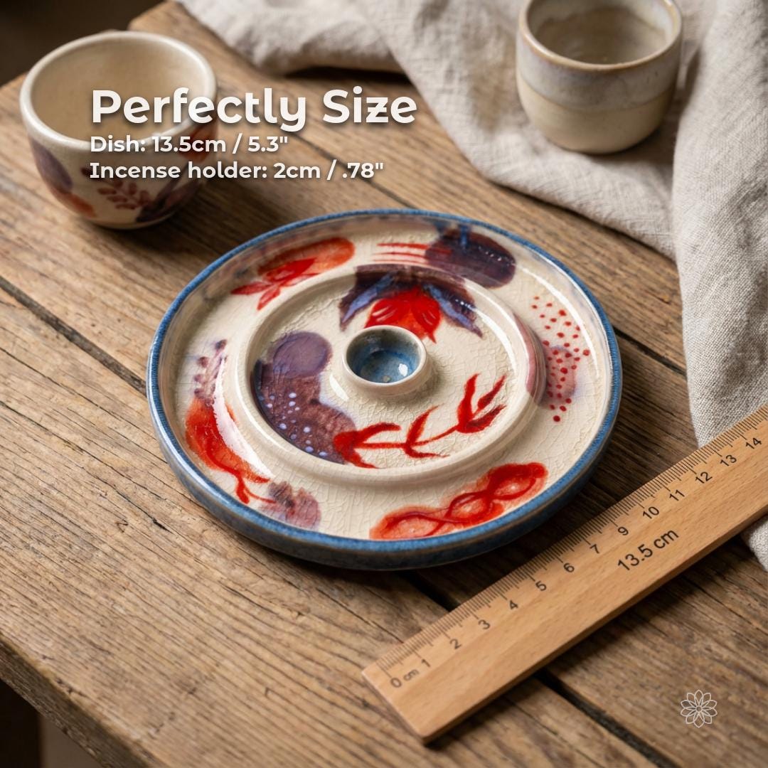 Top-down view of a hand-painted ceramic incense dish showing size and proportions. The dish measures approximately 13.5 cm (5.3 inches) in diameter with a central incense holder opening about 2 cm (0.78 inches). Visible crackle glaze texture, abstract red and purple painted motifs, blue rim, and a wooden ruler placed alongside for scale.