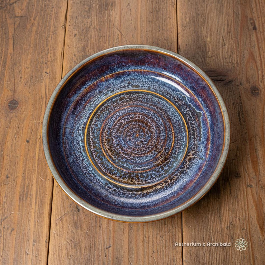 “Stoneware pasta bowl with a deep reactive glaze in blues, indigos, rust, and cream, shown from above on a warm farmhouse wood table. The bowl has a wide rim, gently sloped sides, and a textured spiral pattern. Handmade ceramic piece from the Aetherium × Archibald collection.”