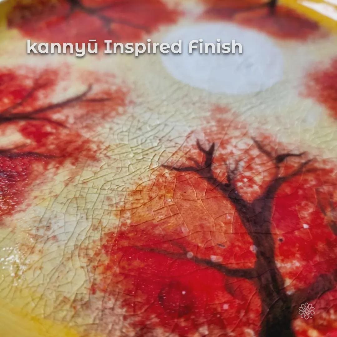 Close-up of a hand-painted ceramic plate surface highlighting kannyū-inspired crackle glaze. Fine natural glaze lines spread across warm cream and yellow tones beneath red tree imagery, emphasizing texture, imperfection, and traditional Japanese pottery influence.