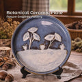 Handmade ceramic dinner plate displayed upright on a black stand, featuring a carved botanical scene with white leaf and plant forms set against a deep blue glaze. The lower portion is softly textured in pale clay tones, creating a horizon-like contrast. Styled on a rustic wooden table with natural textiles and dried botanicals, emphasizing handcrafted detail and calm, nature-inspired design.