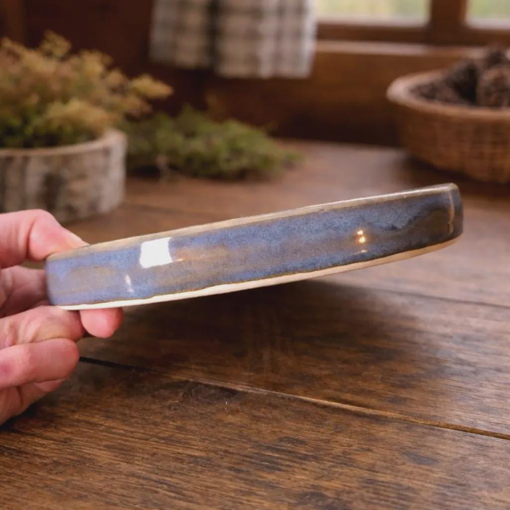 Side profile view of a handmade ceramic plate held in one hand, showing its thickness, rounded rim, and glossy blue glaze. The edge reveals natural clay tones beneath the glaze, emphasizing the plate’s sturdy, wheel-thrown construction and functional design intended for everyday use.