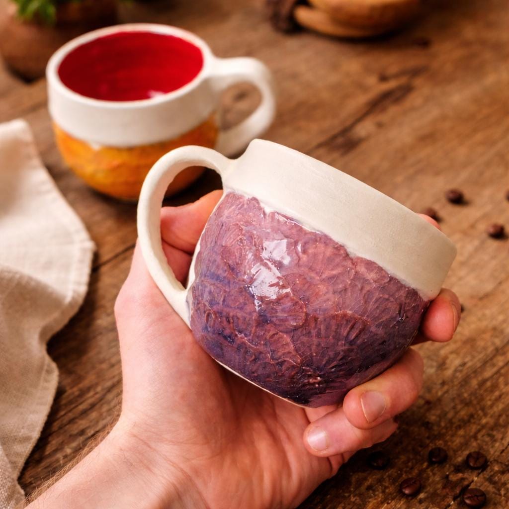 Hand holding a handmade ceramic mug with a deep plum exterior featuring carved, overlapping leaf-like textures. The mug has a thick cream-colored rim, a smooth curved handle, and a dark glazed interior, showing scale and how the mug fits comfortably in the hand.