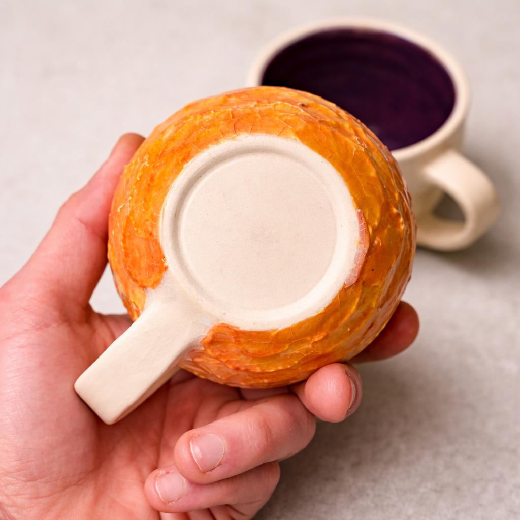 Bottom view of a handmade ceramic mug showing the unglazed foot ring and the transition between the textured orange exterior glaze and the natural clay base. The rounded form and thickness of the ceramic are clearly visible.