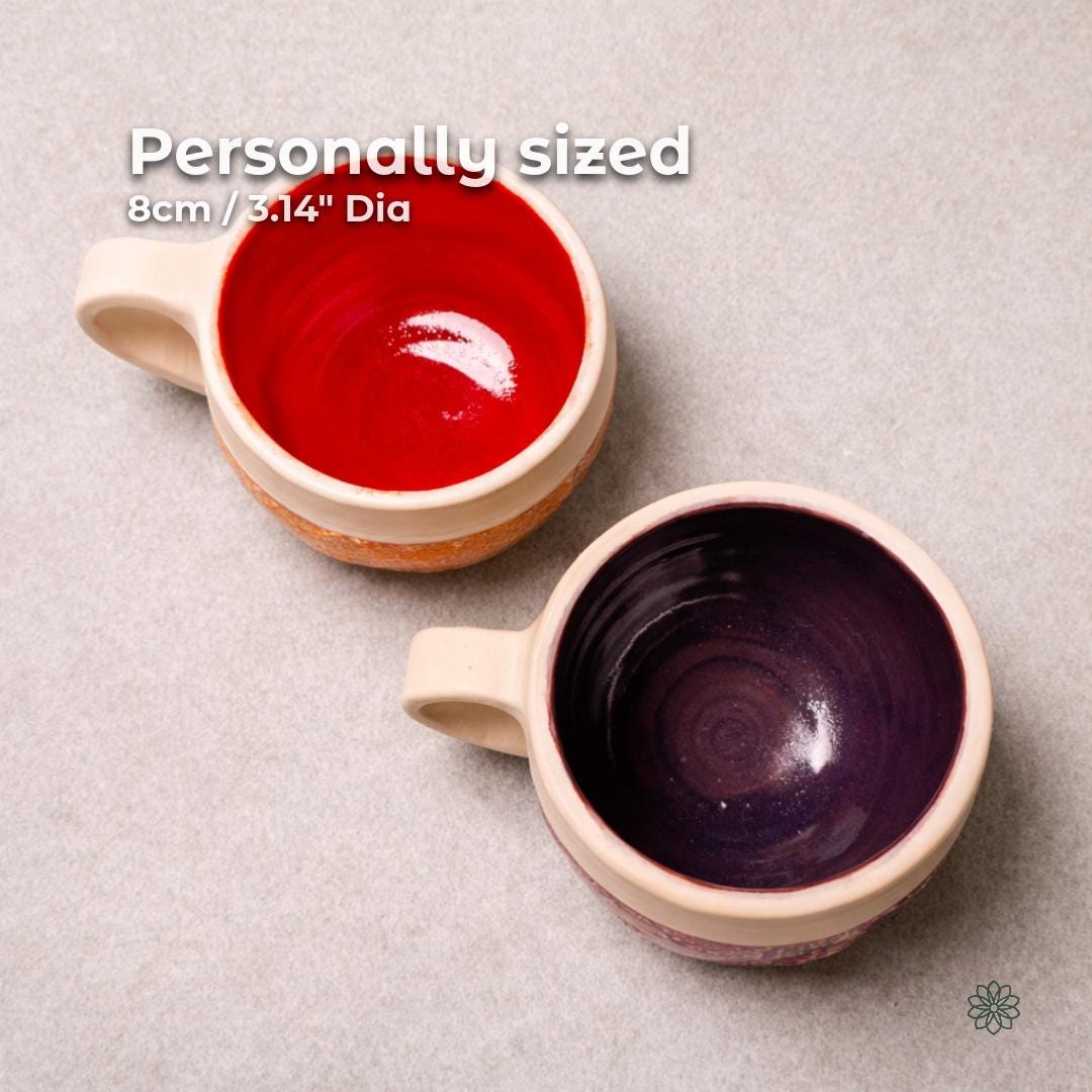 Top-down view of two handmade ceramic mugs showing contrasting interior glazes, one glossy red and one deep plum. The mugs have thick cream-colored rims and rounded handles. Text indicates an opening diameter of approximately 8 centimeters or 3.14 inches.