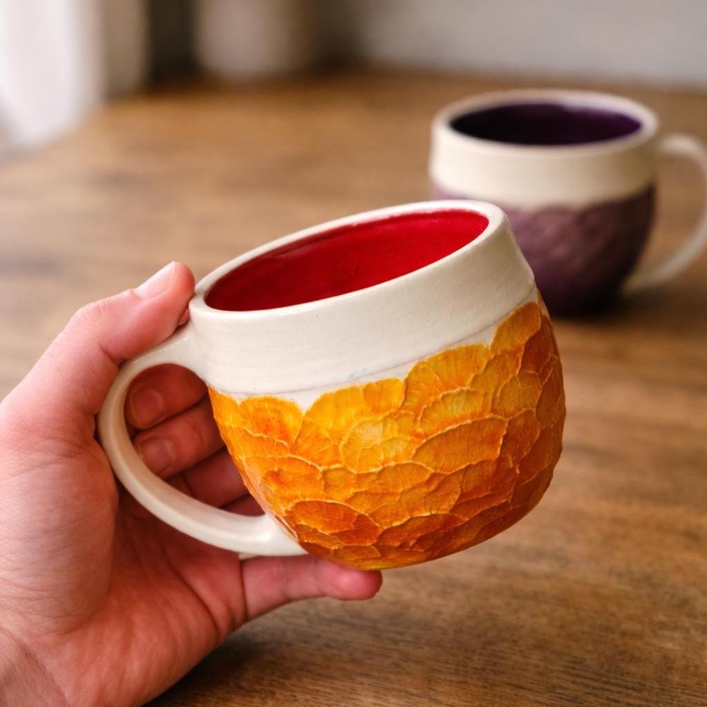 Hand holding a handmade ceramic mug with a warm orange carved exterior and a glossy red interior glaze. The mug features a thick cream-colored rim, rounded handle, and textured surface with layered, organic carving visible across the body.