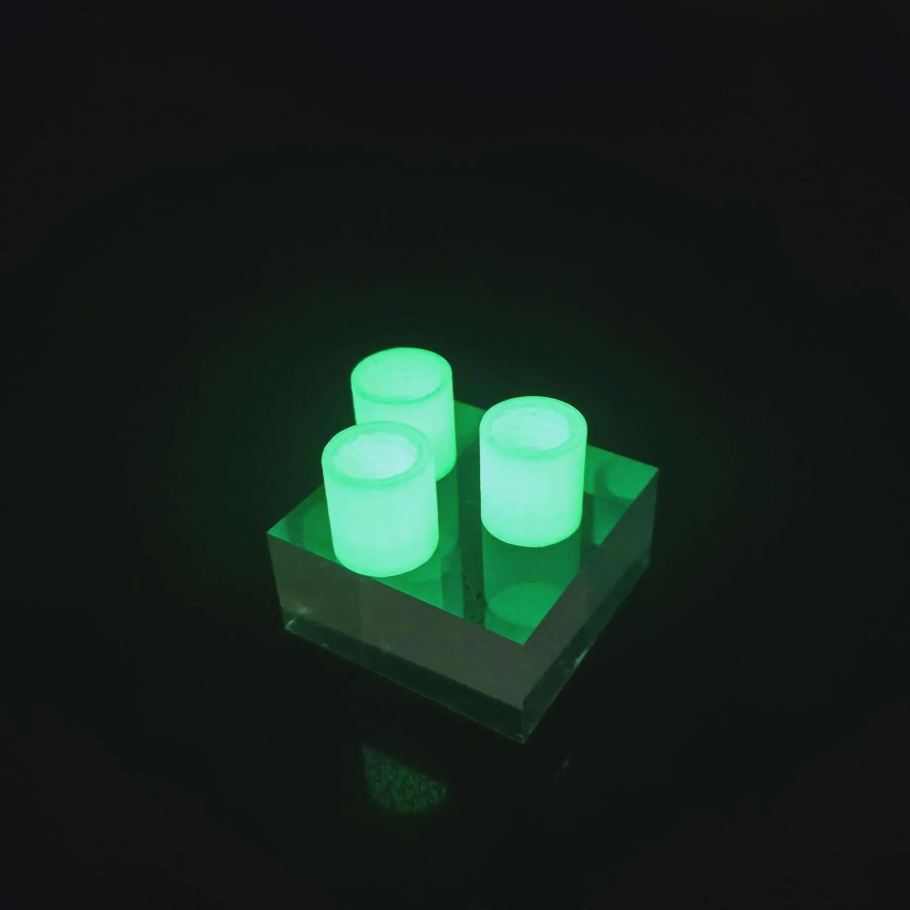 Three glow-in-the-dark toothpaste transfer adapters illuminated in low light on a clear acrylic block. The bright green glow highlights nighttime visibility for campsite or travel use, showing how the adapters can be easily found in dark packs or tents.