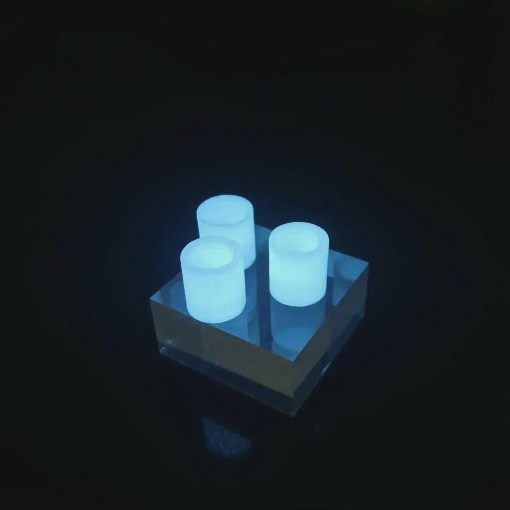 Three glow-in-the-dark toothpaste transfer adapters illuminated in low light on a clear acrylic block. The bright blue glow highlights nighttime visibility for campsite or travel use, showing how the adapters can be easily found in dark packs or tents.