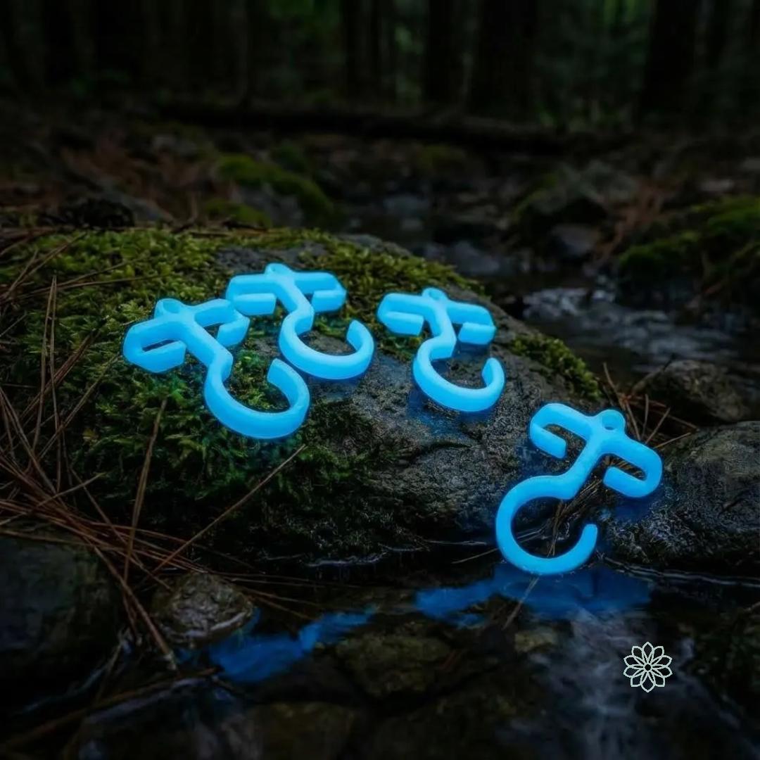 Full-size blue glow-in-the-dark paracord hooks arranged on mossy rocks beside a stream at dusk. Highlights visibility at night, durable bioplastic construction, and utility for camping, hiking, and bushcraft gear organization.