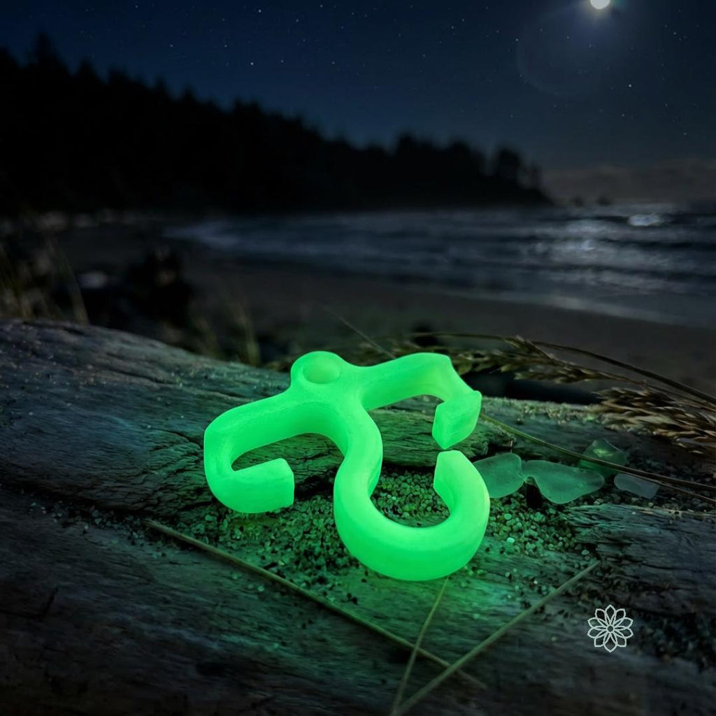 Bright blue glow-in-the-dark mini paracord hook resting on driftwood at a moonlit beach. Soft shoreline and night sky emphasize nighttime visibility for backpackers and campers using ultralight organization tools.
