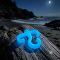 Ultra-bright blue glow mini paracord hook glowing on a driftwood log near the ocean. The night sky and shoreline emphasize high nighttime visibility for hiking or emergency gear use.