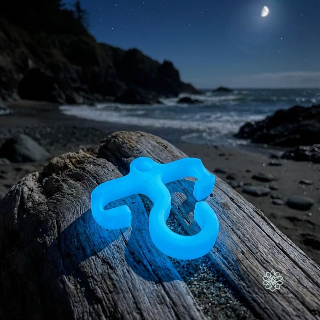 Ultra-bright blue glow mini paracord hook glowing on a driftwood log near the ocean. The night sky and shoreline emphasize high nighttime visibility for hiking or emergency gear use.