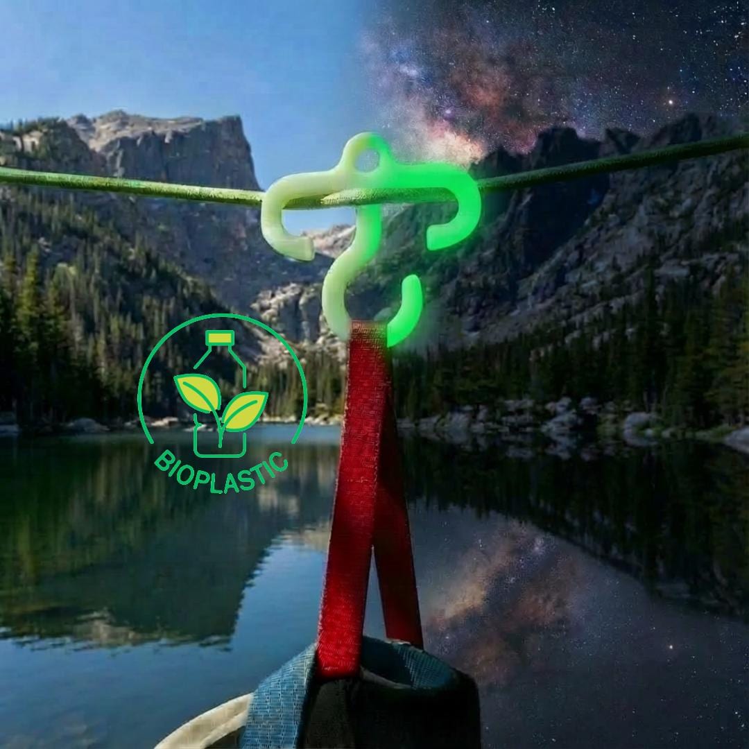 Glow-in-the-dark mini paracord hook hanging from a green line above a mountain lake. The bioplastic camping hook holds a red gear strap, with a split day-and-night sky highlighting its visibility. Designed for ultralight backpacking and overnight camp organization.