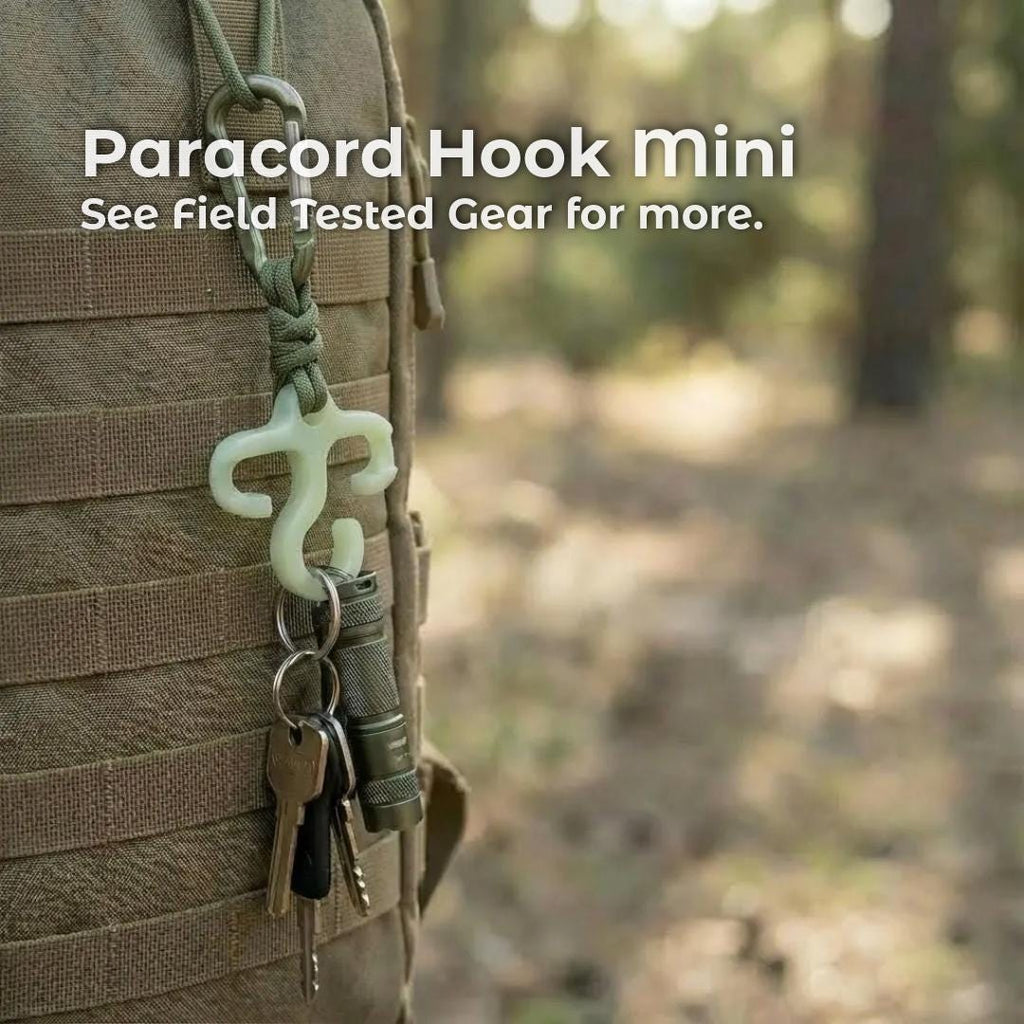 A small glow-in-the-dark paracord hook hangs from green cord on the side of a tan backpack, holding keys and a compact flashlight. The hook is shown in an outdoor forest setting to illustrate its ultralight, field-tested use for backpackers and hikers.