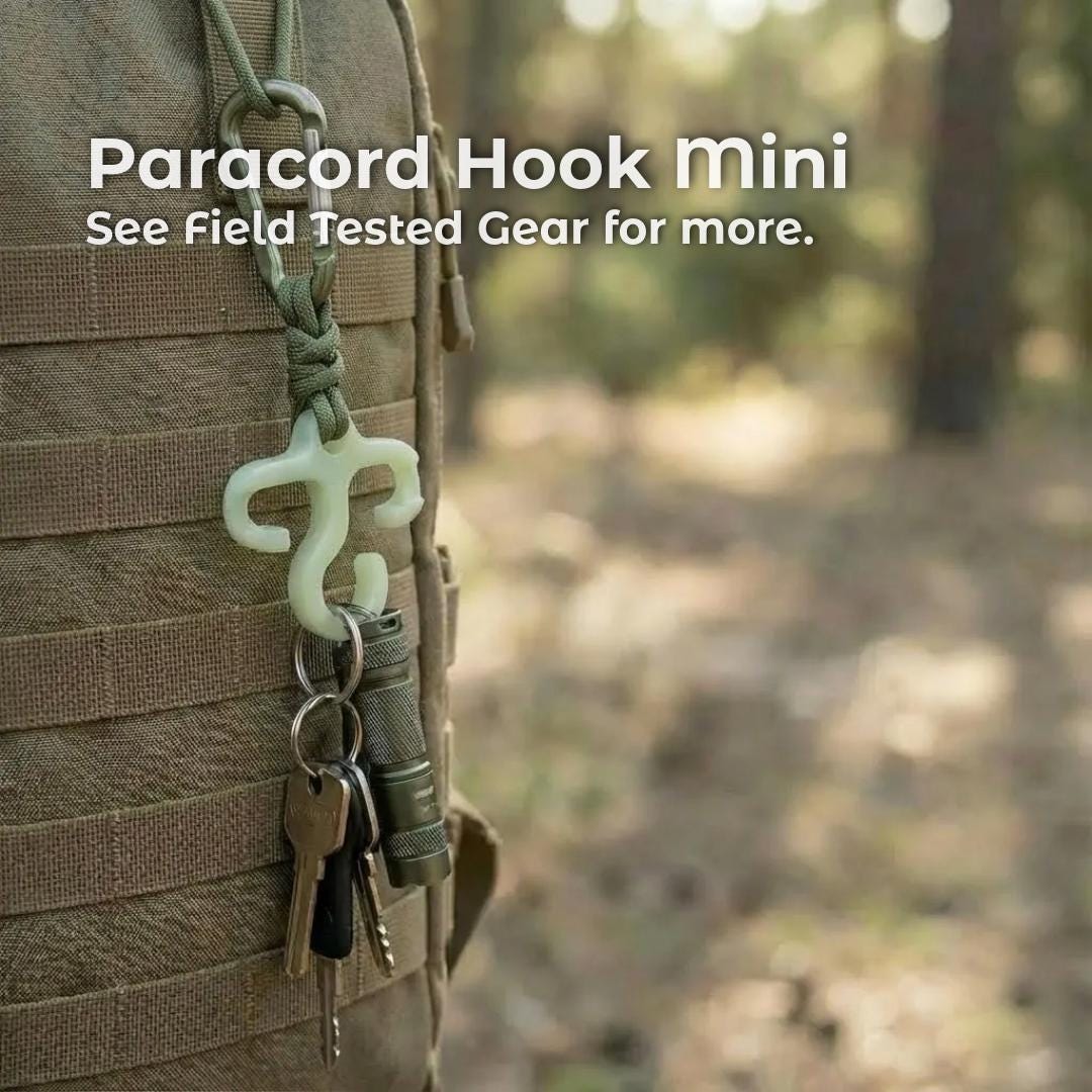 A small glow-in-the-dark paracord hook hangs from green cord on the side of a tan backpack, holding keys and a compact flashlight. The hook is shown in an outdoor forest setting to illustrate its ultralight, field-tested use for backpackers and hikers.