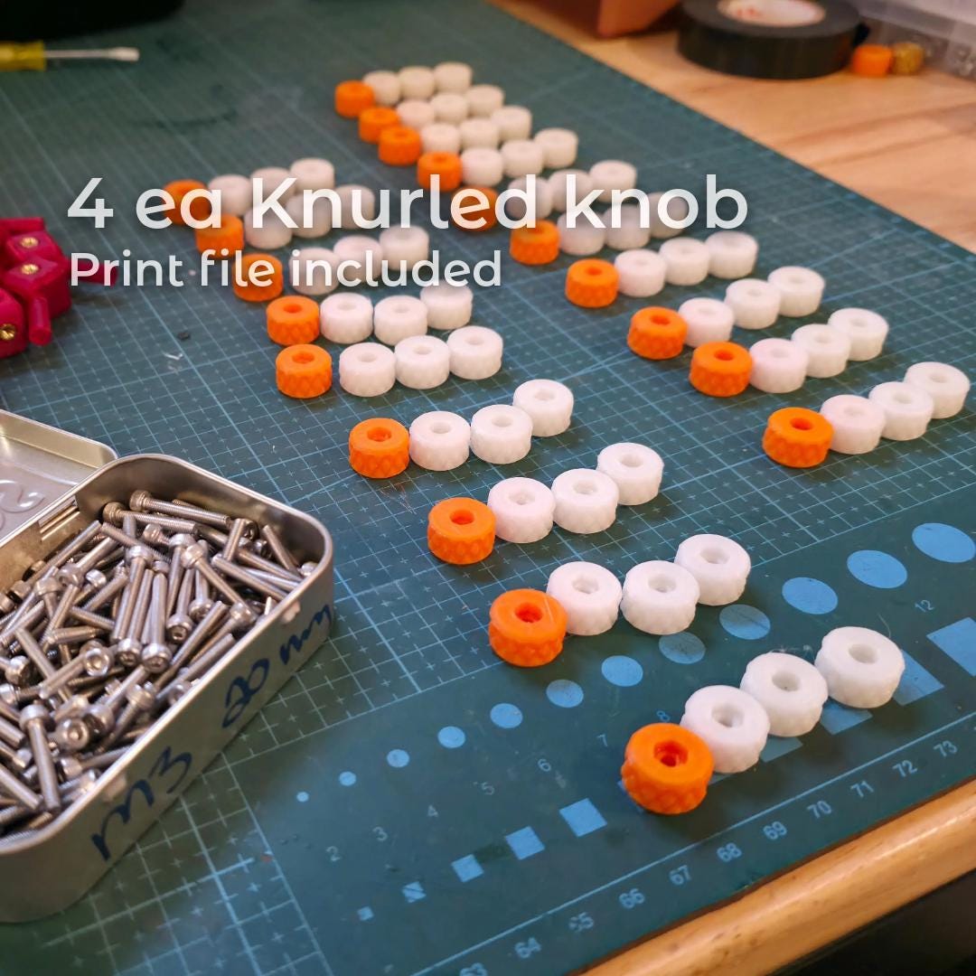 Rows of printed knurled adjustment knobs in white and orange polycarbonate arranged on a cutting mat beside stainless hardware. The image highlights included knob design files that provide tactile control for fine centering adjustments during gemstone alignment.