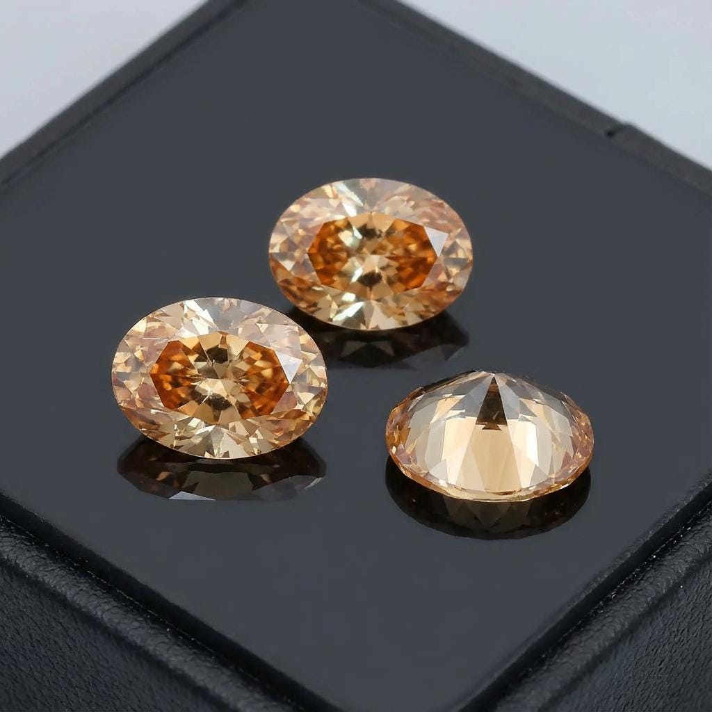 “Three champagne oval CZ gemstones on a reflective black surface. One stone is tilted to show pavilion detail. The gems feature even golden-champagne color, crisp faceting, and calibrated 9×7 mm measurements used in rings, pendants, and bespoke jewelry fabrication.”