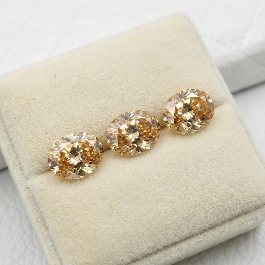 “Three champagne-colored oval CZ gemstones arranged on a cream velvet jewelry pad. The stones show strong brilliance, symmetrical faceting, and even color distribution. Their calibrated 9×7 mm size makes them suitable for jewelers working with premade or custom settings.”