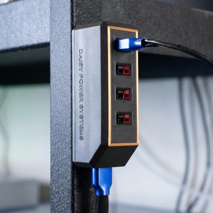 Magnetic Anderson Powerpole DC power distribution block mounted vertically on a steel shelf post, branded Daisy Power Systems. The compact rectangular housing features four red and black Anderson Powerpole output ports on the front and a single Anderson input port on the bottom, with blue power cables connected. Designed for universal 12V DC power distribution in workshops, off-grid systems, campers, and maker setups.