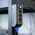 Magnetic Anderson Powerpole DC power distribution block mounted vertically on a steel shelf post, branded Daisy Power Systems. The compact rectangular housing features four red and black Anderson Powerpole output ports on the front and a single Anderson input port on the bottom, with blue power cables connected. Designed for universal 12V DC power distribution in workshops, off-grid systems, campers, and maker setups.