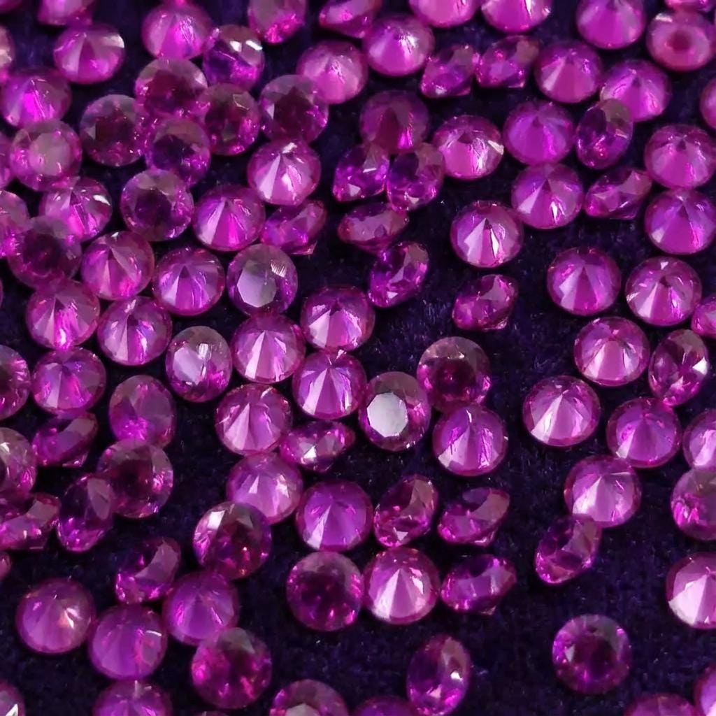 Close-up of multiple lab grown pink sapphire accent stones, 2 mm round cut, clustered on dark fabric. Vivid pink color and uniform faceting shown across many stones, ideal for jewelers needing matching embellishment stones for bands, halos, or pavé work.