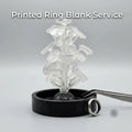 Clear PolyCast PVB 3D printed ring blanks arranged on a sprue tree, standing upright in a black casting base. Multiple oval, round, and rectangular ring blank shapes are visible, showing smooth surfaces and uniform wall thickness. A finished silver ring rests beside the base, suggesting the transition from printed casting pattern to completed jewelry. Neutral studio background with soft lighting emphasizes material clarity and form.
