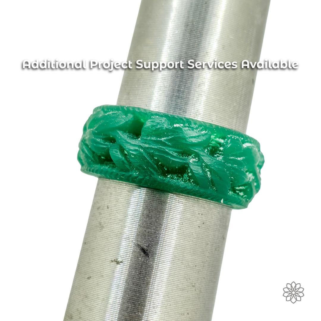 A carved green wax ring blank mounted with a machined leaf pattern, on a metal mandrel, showing textured surface detail and unfinished edges prior to casting. The ring appears mid-process, illustrating a traditional wax stage used before metal casting or finishing. Text on the image reads “Additional Project Support Services Available,” signaling optional fabrication or production assistance beyond the ring blank itself.