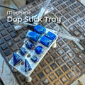 Magnetic dop stick tray holding polished blue lapis lazuli cabochons and geometric stones on metal dops. The white bioplastic tray organizes multiple stones upright during lapidary cutting and faceting, shown on a textured work surface with syringes and tools in the background. Designed for lapidary artists using dop sticks, faceting machines, and organized stone-setting workflows.