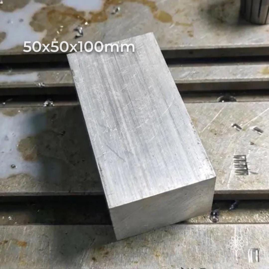 Raw 50×50×50 mm 6061-T6 aluminum cube resting on a CNC machine table with coolant residue and metal shavings. Light tool lines on the face. High-quality machining stock for milling, drilling, lathe work, custom components, and metal prototyping.