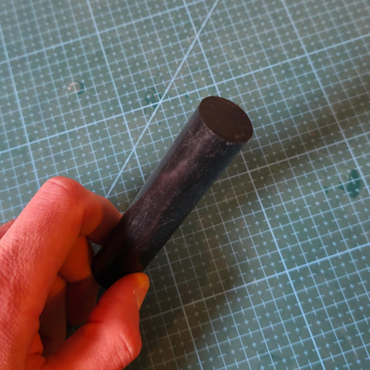 Close-up of a black Delrin acetal rod held in hand over a cutting mat, showing the solid round cross section and matte surface. Engineering plastic rod stock suitable for lathe turning, CNC machining, and fabrication of low-friction mechanical parts.