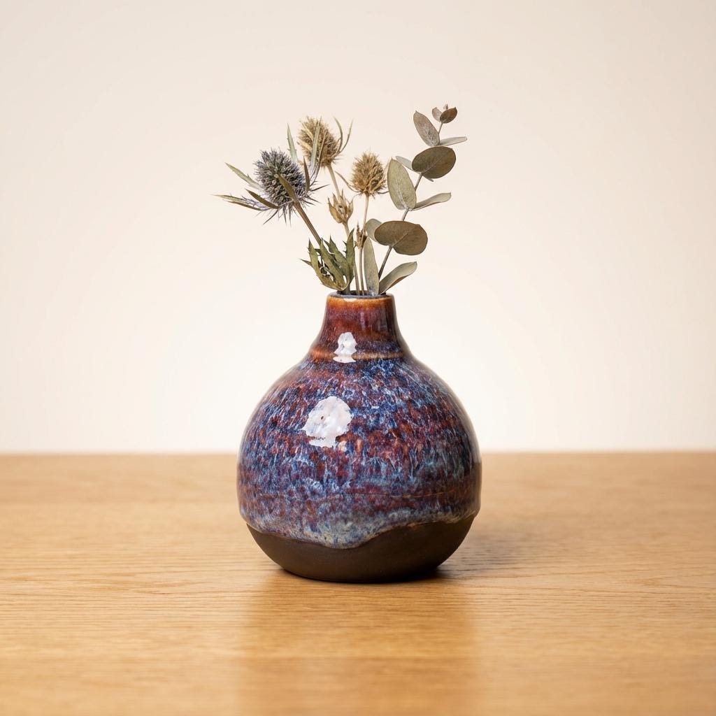 Handmade ceramic art vase with a rounded, grounded form and narrow neck, holding a minimal arrangement of dried botanical stems. The reactive glaze displays layered blue, violet, and rust coloration with soft white interruptions, transitioning to a darker lower band. Photographed on a wooden surface against a light background to show overall silhouette and scale.