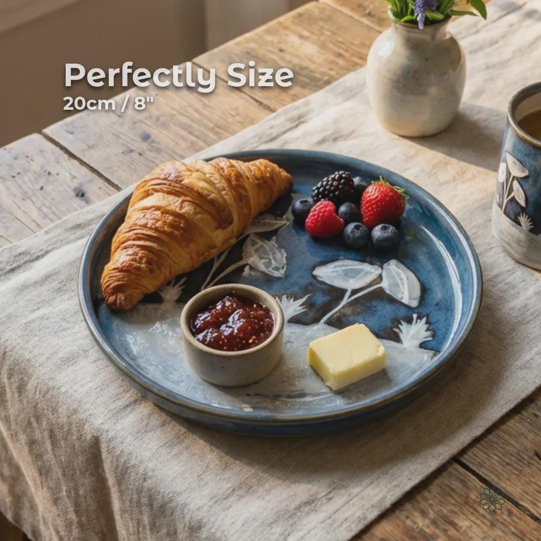 Handmade ceramic plate in use on a wooden table, holding a croissant, berries, butter, and jam. The plate’s blue glaze and carved botanical design are visible beneath the food, showing scale and everyday functionality. White text on the image indicates the plate measures approximately 20 cm or 8 inches in diameter.