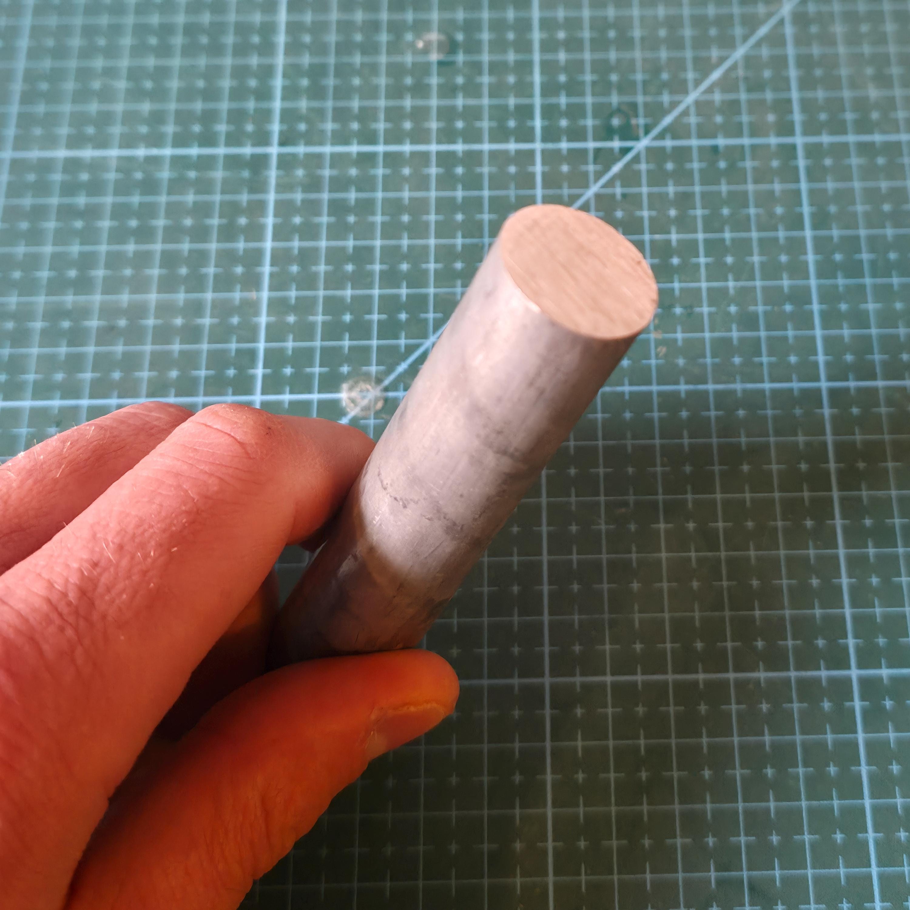Close-up of a hand holding a 7075 aluminum round bar over a gridded cutting mat, showing real-world scale, surface finish, and material texture. Solid aluminum rod stock suitable for CNC machining, lathe turning, prototypes, fixtures, and small-batch mechanical parts.