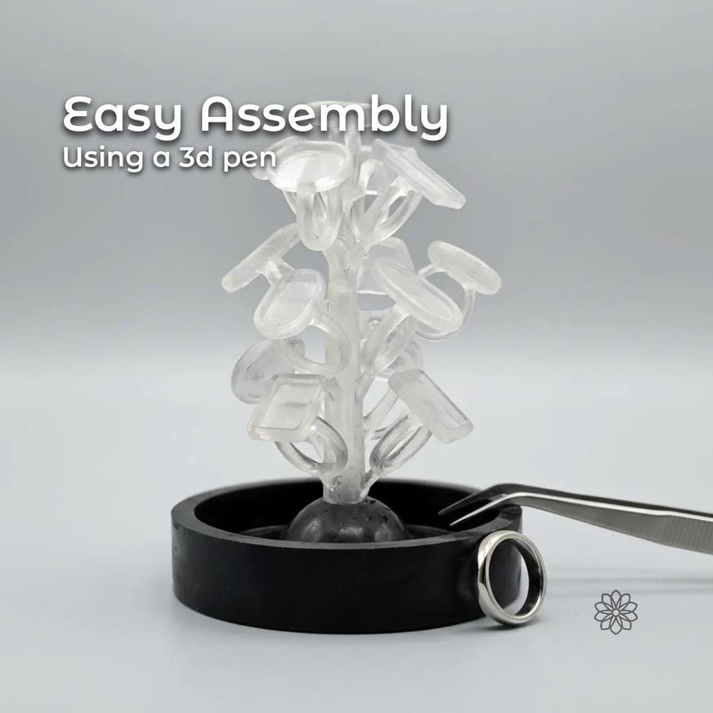 Clear resin sprue tower holding multiple ring blank wax patterns arranged vertically, demonstrating easy assembly for lost wax casting. The sprue tree stands in a black rubber casting base on a neutral gray background. Metal tweezers and a finished silver ring sit beside the base, showing scale and use. Overlay text reads “Easy Assembly Using a 3D Pen,” highlighting the simple spruing process for jewelry makers.