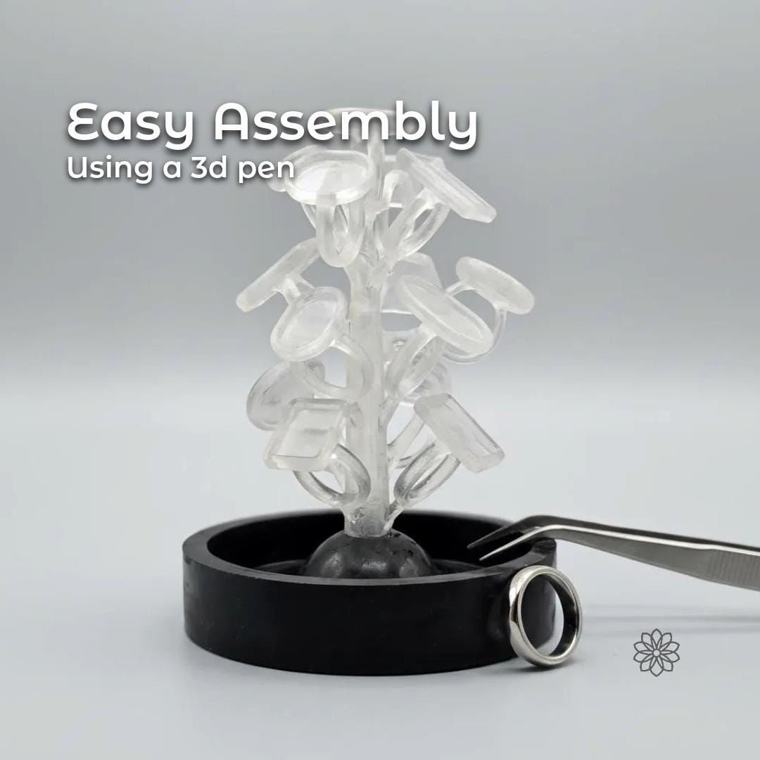 Clear resin sprue tower holding multiple ring blank wax patterns arranged vertically, demonstrating easy assembly for lost wax casting. The sprue tree stands in a black rubber casting base on a neutral gray background. Metal tweezers and a finished silver ring sit beside the base, showing scale and use. Overlay text reads “Easy Assembly Using a 3D Pen,” highlighting the simple spruing process for jewelry makers.