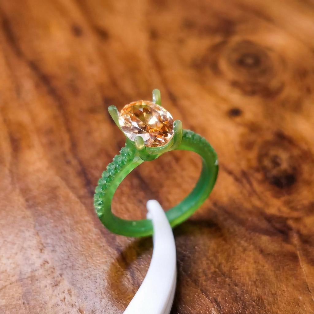 A translucent green resin test ring holding a faceted round gemstone in a prong setting, shown resting on a wooden work surface. A white tool gently touches the ring near the stone, illustrating test fitting and inspection before casting. The image highlights careful handling, stone protection, and fit verification during the jewelry development process, prior to metal casting or final finishing.