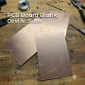 “Three double-sided copper-clad PCB boards arranged on a workbench surrounded by electronics tools, resistors, cutters, and solder. Each board has copper laminate on both faces, designed for CNC milling, DIY circuit etching, dual-layer prototypes, and custom electronics projects.”