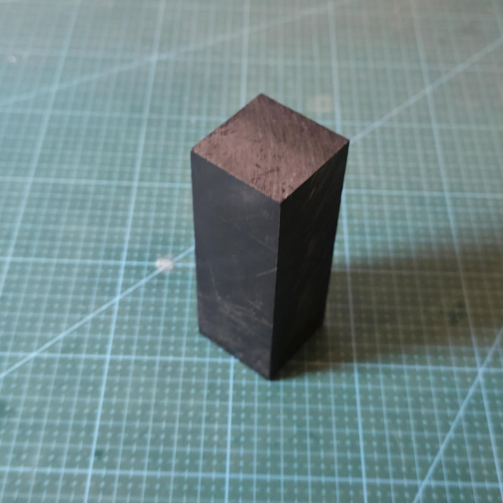 Black Delrin square block standing upright on a cutting mat, showing the square end face and uniform material structure. The solid acetal plastic construction is clearly visible, illustrating consistent density and clean cut faces ideal for CNC machining, jig making, and precision mechanical components.