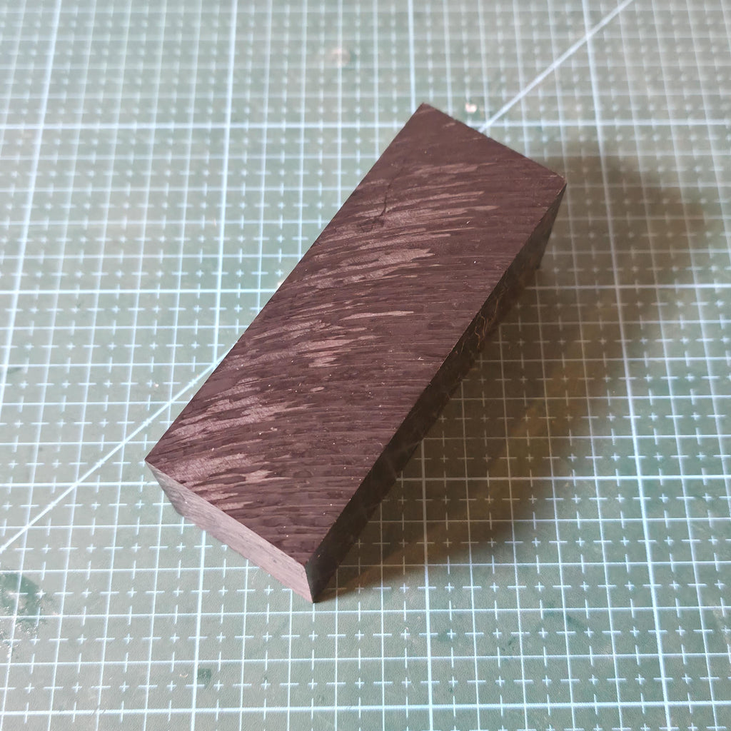 Black Delrin square stock block shown on a gridded cutting mat at an angle, displaying the long face and sharp edges of the acetal material. Surface texture and machining marks are visible, emphasizing dimensional stability, low friction plastic properties, and suitability for CNC milling, drilling, and fixture fabrication.