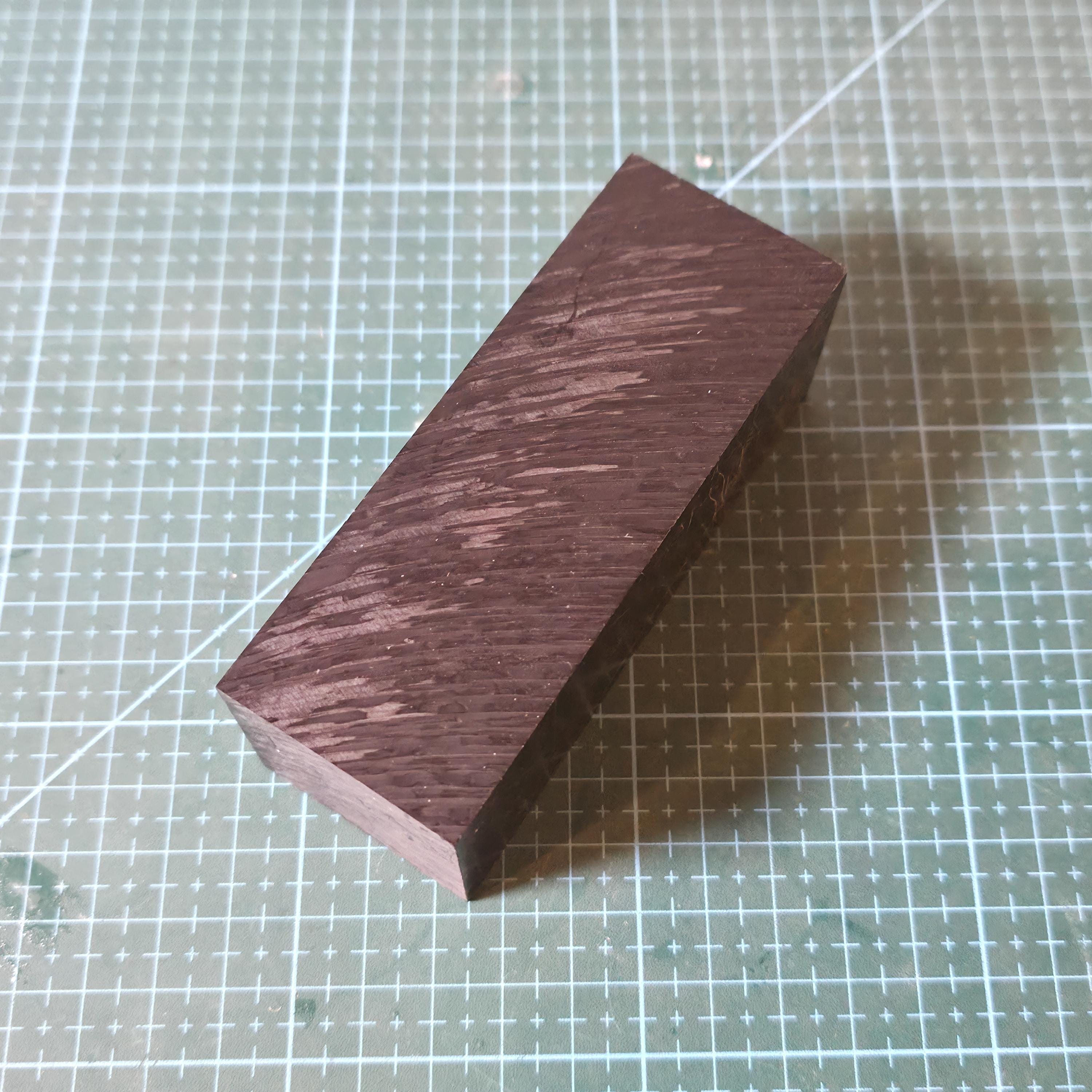 Black Delrin square stock block shown on a gridded cutting mat at an angle, displaying the long face and sharp edges of the acetal material. Surface texture and machining marks are visible, emphasizing dimensional stability, low friction plastic properties, and suitability for CNC milling, drilling, and fixture fabrication.
