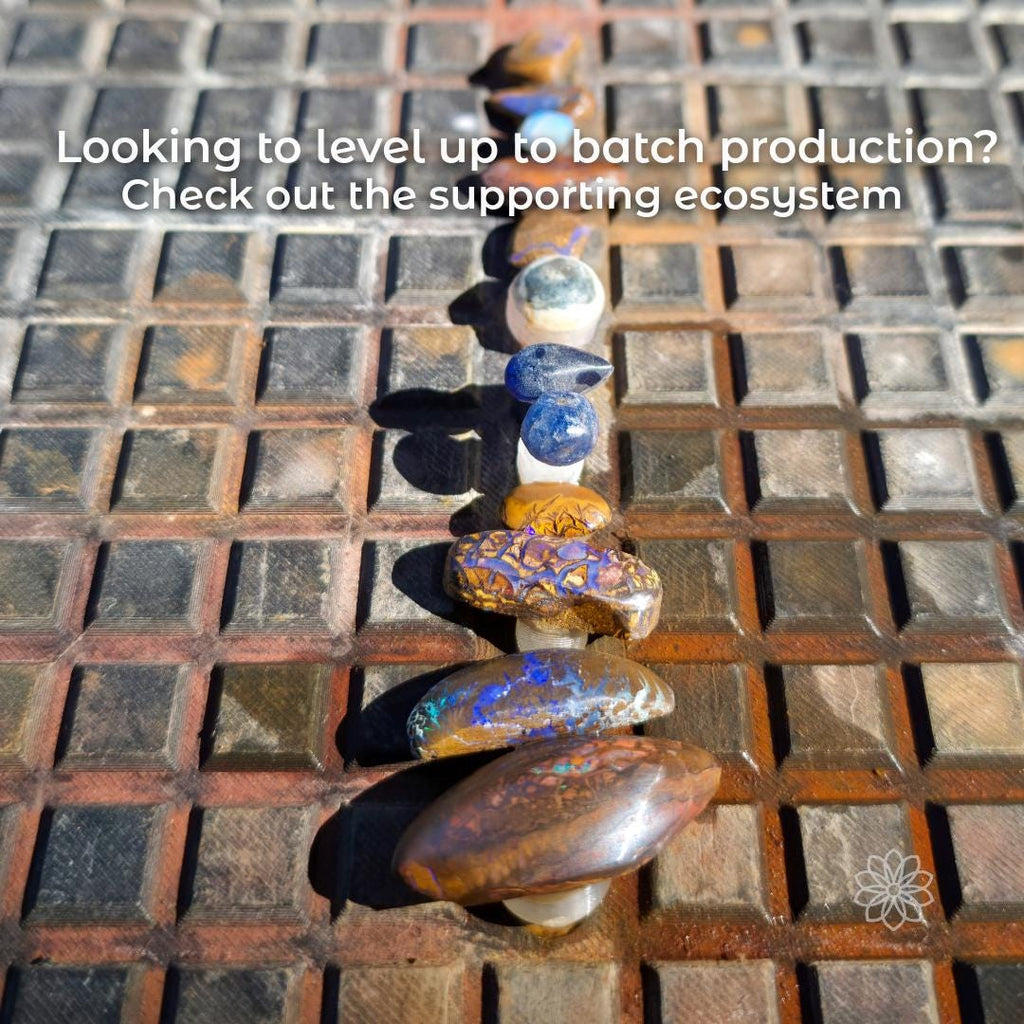 Row of gemstone dops arranged in sequence on a textured work surface, with overlaid text reading “Looking to level up to batch production? Check out the supporting ecosystem,” illustrating how the magnetic dop stick tray supports staged, repeatable batch workflows within a modular lapidary tool system.
